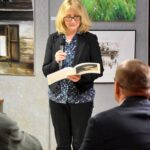Na holu biblioteki Joanna Błońska czyta tekst z albumu za nią wiszą obrazy przedstawiające chaty kryte strzechą.