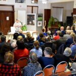 Pani Teresa Nazarczuk podczas prelekcji o tradycjach kulinarnych Podlasia, wygłaszanej w ramach Festiwalu Literatury Kulinarnej w sokołowskiej Bibliotece. Na sali siedzi kilkadziesiąt osób zasłuchanych w opowieść, w tle widoczne są zdjęcia, obrazy i dekoracyjne paprocie.