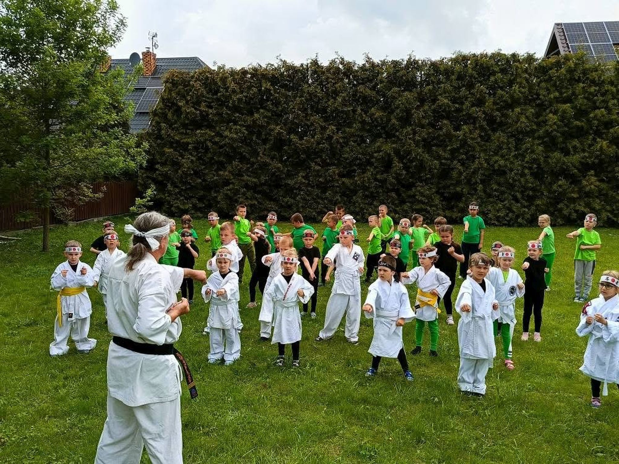 Grupa około trzydziestu przedszkolaków na zewnątrz na trawniku. Dzieci stoją w kilku rządach. Dzieci z przodu ubrane są w białe japońskie kimona. Dzieci z tyłu ubrane są w czarne i zielone stroje. Wszystkie dzieci na czołach mają założone opaski z japońskimi wzorami. Dzieci powtarzają ćwiczenia wykonywane przez trenera karate.