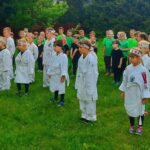 Grupa około trzydziestu przedszkolaków na zewnątrz na trawniku. Dzieci stoją w kilku rządach. Dzieci z przodu ubrane są w białe japońskie kimona. Dzieci z tyłu ubrane są w czarne i zielone stroje. Wszystkie dzieci na czołach mają założone opaski z japońskimi wzorami. Dzieci stoją na baczność.