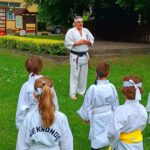 Na środku stoi trener karate, ma na sobie białe kimono oraz czarny pas - to symbol mistrzostwa i wysokiego stopnia zaawansowania w tej sztuce walki. Przed nim stoi czwórka dzieci w kimono. Jedno z nich ma żółty pas, który świadczy o kolejnym stopniu doświadczenia.