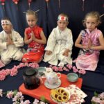 Czworo dzieci ubranych w kimona i stroje karate siedzi przy stoliku przykrytym czerwoną tkaniną, na której ustawione są japońska zastawa do herbaty i ciasteczka. Dzieci mają złożone ręce, a wokół rozłożone są różowe kwiaty wiśni, tworząc nastrój japońskiej ceremonii.