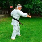 Trener karat w białym kimono z czarnym pasem. Stoi boso na trawniku i wykonuje ćwiczenia.