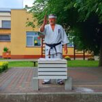 Trener karat w białym kimono z czarnym pasem oraz białą opaską na czole. Stoi na kostce. Przed nim leży pięć bloczków przełożonych drewnianymi listewkami. Jest to pokaz rozbijania bloczków gołą ręką.