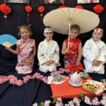 Czworo dzieci ubranych w kimona i stroje karate siedzi na tle dekoracji z japońskimi motywami. Dwie dziewczynki trzymają wachlarz i parasol, a dwóch chłopców w białych strojach karate i opaskach siedzi obok. Przed nimi ustawiony jest stolik z naczyniami do herbaty i ciasteczkami, otoczony kwiatami wiśni.