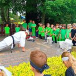 Trener karate w białym kimono z czarnym pasem wykonuje pompki na opierając się na pięściach. Wokół zebranie dzieci z przedszkola. Dzieci mają na sobie białe kimona, czarne stroje oraz zielone stroje.