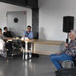 Autor Andrzej Ziemiański siedzi na krześle zwrócony w kierunku kilku osób siedzących po przeciwnej stronie, kobieta w sukience - dyrektor biblioteki Małgorzata Iwańska-Kania stoi za nim obok kolumny.