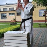 Trener karat w białym kimono z czarnym pasem oraz białą opaską na czole. Stoi na kostce. Przed nim leży pięć bloczków przełożonych drewnianymi listewkami. Jest to pokaz rozbijania bloczków gołą ręką.