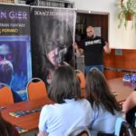 Leszek Bigos stoi przed plakatami promującymi „Salon gier” i „Smoczy szlak. Wejście”. Widoczne jest także kilku uczestników siedzących przy stole, słuchających autora.