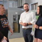 Trzy osoby stoją i rozmawiają w pomieszczeniu biblioteki, dyrektor biblioteki Małgorzata Iwańska-Kania trzyma obraz, który otrzymała od uczestników kursu.