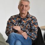 Autor Andrzej Ziemiański w koszuli w kolorowe kwiatowe wzory siedzi na krześle, trzyma w dłoni mikrofon i patrzy w stronę fotografa.