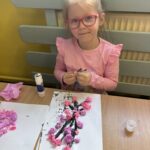 Dziewczynka w różowych okularach siedzi przy stoliku i tworzy pracę plastyczną z motywem kwitnącej wiśni, wykorzystując różowe i fioletowe kuleczki z bibuły.