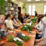 Grupa uczestników warsztatów w bibliotece przygotowuje kadzidła słowiańskie z ziół ułożonych na stole.