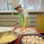 Dziecko w zielonym fartuszku rozwałkowuje ciasto na pierogi, obok tacka z przygotowanymi pierogami oraz miska z farszem serowym.