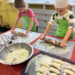 Dziewczynka w różowej koszulce i chłopiec w czapce kucharskiej rozwałkowują ciasto na pierogi, obok tacka z gotowymi pierogami i miska z farszem.
