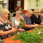 Grupa seniorów w bibliotece wykonuje kadzidła słowiańskie, wiążąc zioła podczas warsztatów.