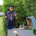 Mężczyzna w czapce i okularach opowiada o pszczelarstwie na tle zielonych krzewów.
