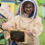 Uśmiechnięta kobieta w stroju pszczelarskim trzyma w dłoniach ramkę z pszczołami.