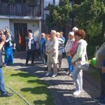 Grupa osób słucha mężczyzny prowadzącego prelekcję na świeżym powietrzu, stojąc na kostce brukowej przy budynku, w słoneczny dzień.
