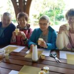 Czworo seniorów siedzi przy stole na świeżym powietrzu, uśmiechając się i trzymając własnoręcznie wykonane świece z wosku pszczelego oraz słoiczki miodu.