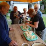 Grupa seniorów stoi wokół stołu pod żółtym namiotem, oglądając i fotografując własnoręcznie wykonane świece z wosku pszczelego ustawione na tackach.