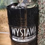 Czarna beczka z napisem „Wystawa starych motocykli, Drohiczyn nad Bugiem” z hełmem, czapką moro i goglami leżącymi na wierzchu.