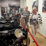 Dwie seniorki spacerują wzdłuż wystawy starych motocykli w muzeum, po lewej stronie widoczne rzędy zabytkowych motocykli oddzielonych czerwoną taśmą.