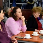 Słuchacze podczas spotkania z Katarzyną Nowosielską siedzą przy stolikach. Na stolikach filiżanki na spodkach.