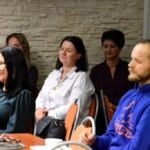 Słuchacze podczas spotkania z Katarzyną Nowosielską siedzą przy stolikach. Na stolikach filiżanki na spodkach. Przy jednym ze stolików siedzi dyrektor biblioteki - Małgorzata Iwańska Kania.