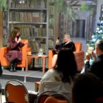 Dwie kobiety siedzą na fotelach. Po lewej stronie Katarzyna Nowosielska, po prawej Bożena Mrozowska-Gozdołek. Kobiety prowadzą rozmowę przez mikrofon. Obok nich z prawej strony stoi udekorowana na biało choinka. Za nimi wielki baner ze zdjęciem regałów z książkami z biblioteki. Na pierwszy planie przed paniami siedzi widownia.