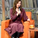 Autorka - Katarzyna Nowosielska siedzi na fotelu, trzyma w ręku mikrofon. Obok niej niski stolik ze spodkiem i kubkiem.