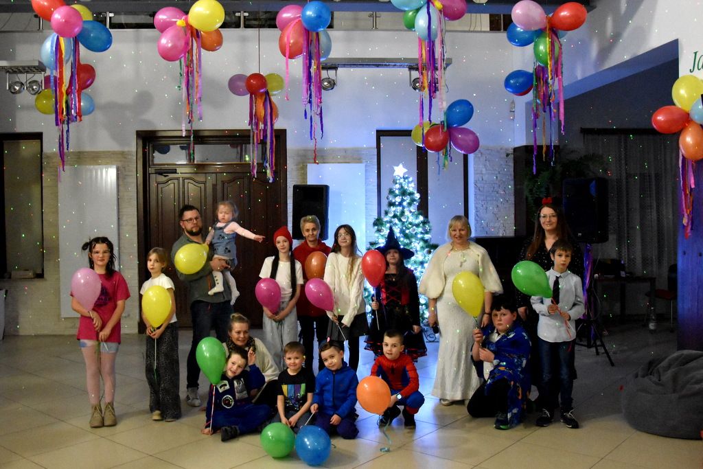 Dzieci i dorośli podczas balu karnawałowego w bibliotece, z kolorowymi balonami.