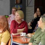 Publiczność podczas spotkania w bibliotece siedzi przy stolikach.