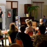 Szeroki kadr, na pierwszym planie siedzi publiczność przy stolikach. Na drugim planie młoda kobieta stoi na scenie, trzyma w ręku mikrofon i śpiewa.