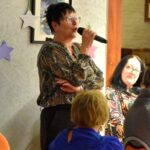 Kobieta z publiczności stoi przy stoliki. Trzyma w ręku mikrofon i przemawia.