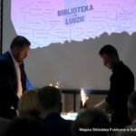 Tort z odpalonymi świecami za nim na ekranie wyświetlana napis biblioteka to ludzie a wokół niego wypisani są wszyscy pracownicy biblioteki miejskiej na przestrzeni 80 lat.