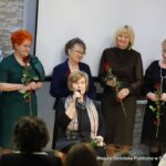 Wąski kadr. Byli pracownicy biblioteki trzymają czerwone róże otrzymane od obecnej dyrektor Małgorzaty Iwańskiej-Kania. W centrum siedzi Wanda Wierzchowska - dyrektor biblioteki w dawnych czasach, za nią stoją m.in. Hanna Lecyk - poprzednia dyrektor biblioteki oraz Bogumiła Korbuszewska - pełniąca obowiązku dyrektora biblioteki.
