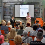 Wiesława Kwiek przemawia do zgromadzonej publiczności podczas spotkania autorskiego w bibliotece. Obok niej przy instrumencie siedzi młody mężczyzna, a na ekranie wyświetlony jest plakat wydarzenia z datą 19 czerwca 2026 roku.