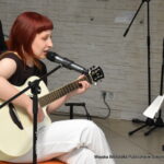 Liwia Długosz śpiewa do mikrofonu i gra na gitarze akustycznej podczas wieczoru autorskiego Wiesławy Kwiek. Przed nią stoi pulpit z nutami.